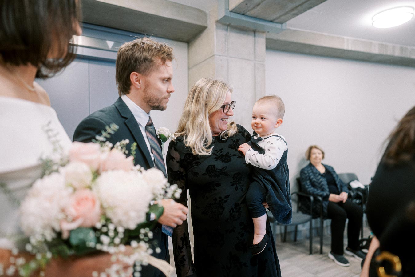 A joyful wedding scene with a woman holding a smiling baby, surrounded by guests in formal attire, with a focus on a nearby bouquet of pink and white flowers.