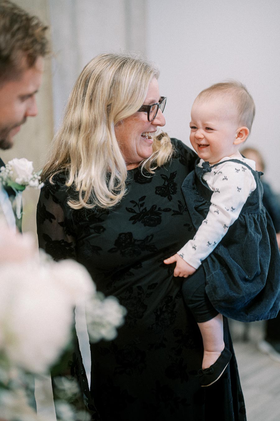 A joyful grandmother holding her smiling grandchild, both dressed in elegant attire at a family gathering.