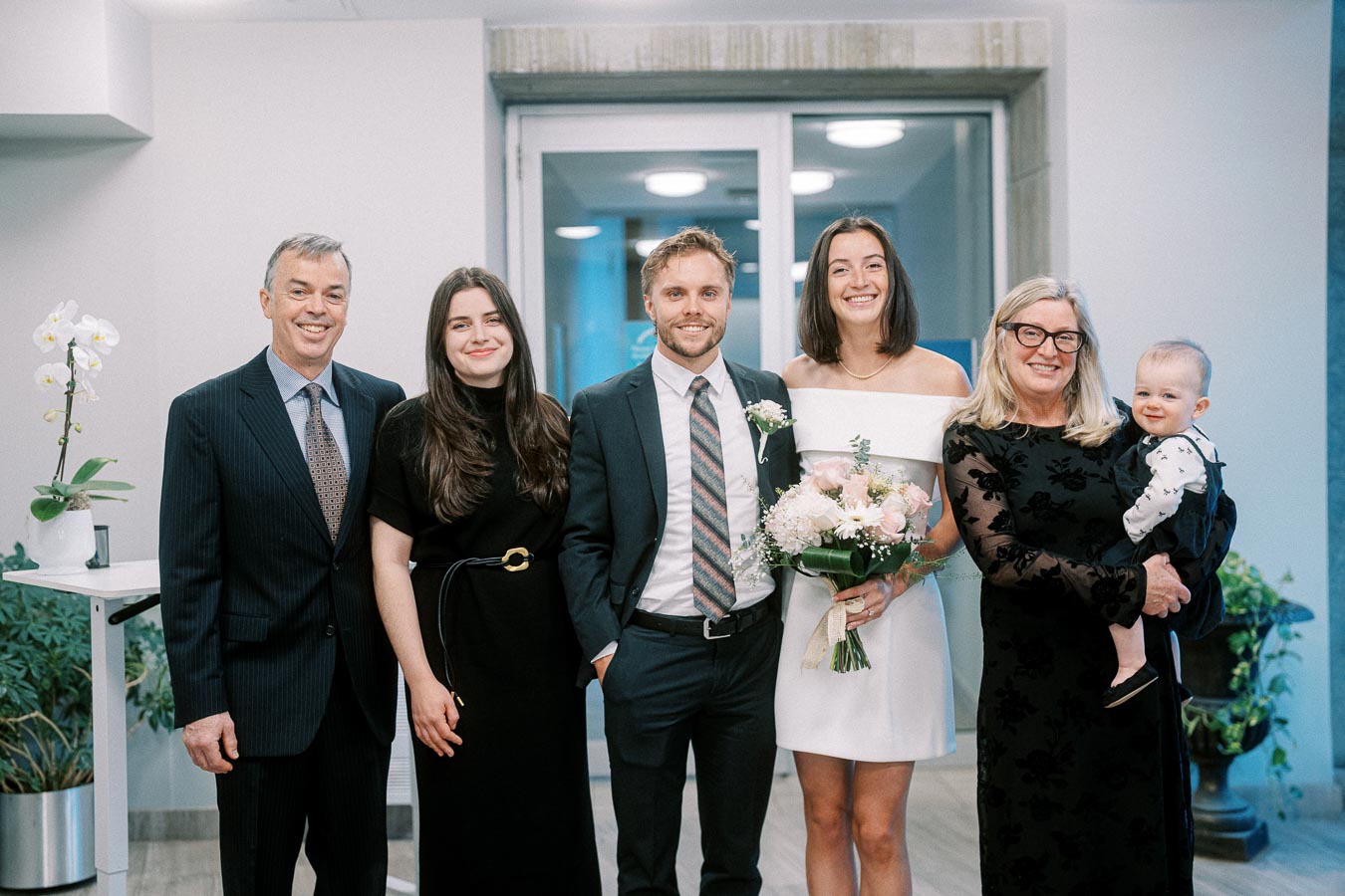 A group of six people, including a couple in formal attire, surrounded by family, posing indoors for a celebration. One person is holding a bouquet of pink and white flowers, and a woman is holding a baby.