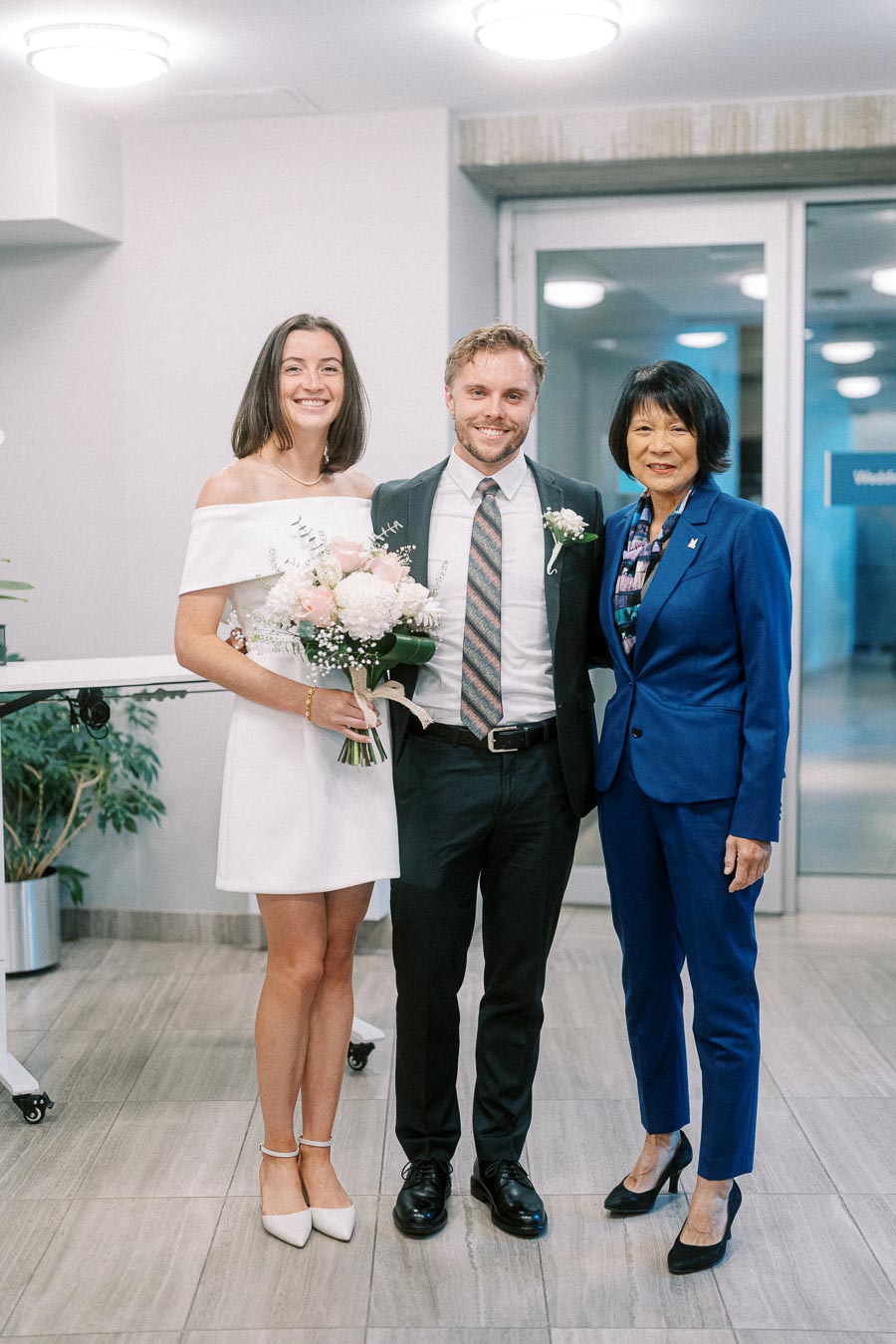 A newlywed couple posing happily with a woman in a blue suit, indoors, the bride holding a bouquet of white and pink flowers.