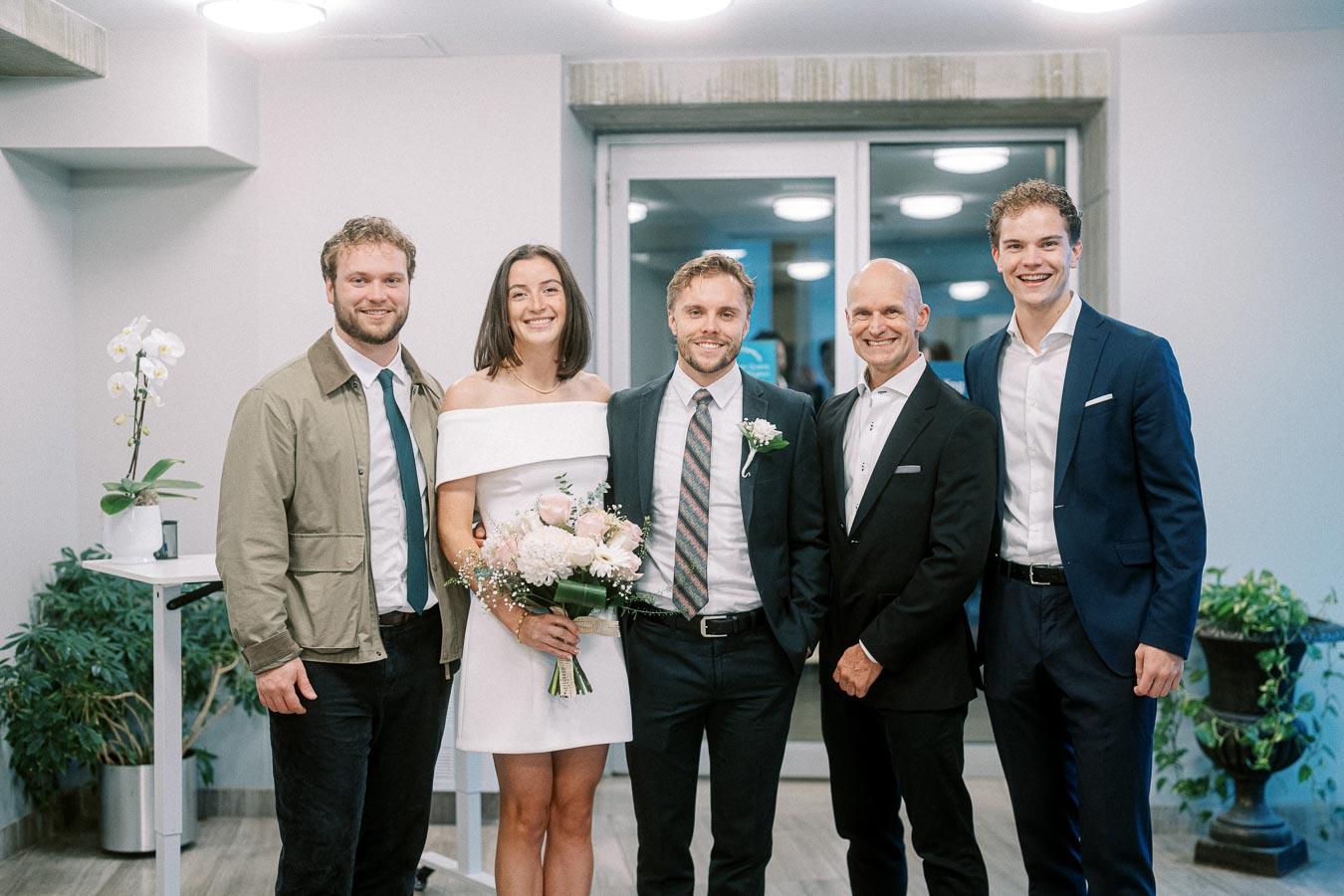 A happy group of people dressed in formal attire, with a woman in a white dress holding a bouquet of flowers, posing indoors.