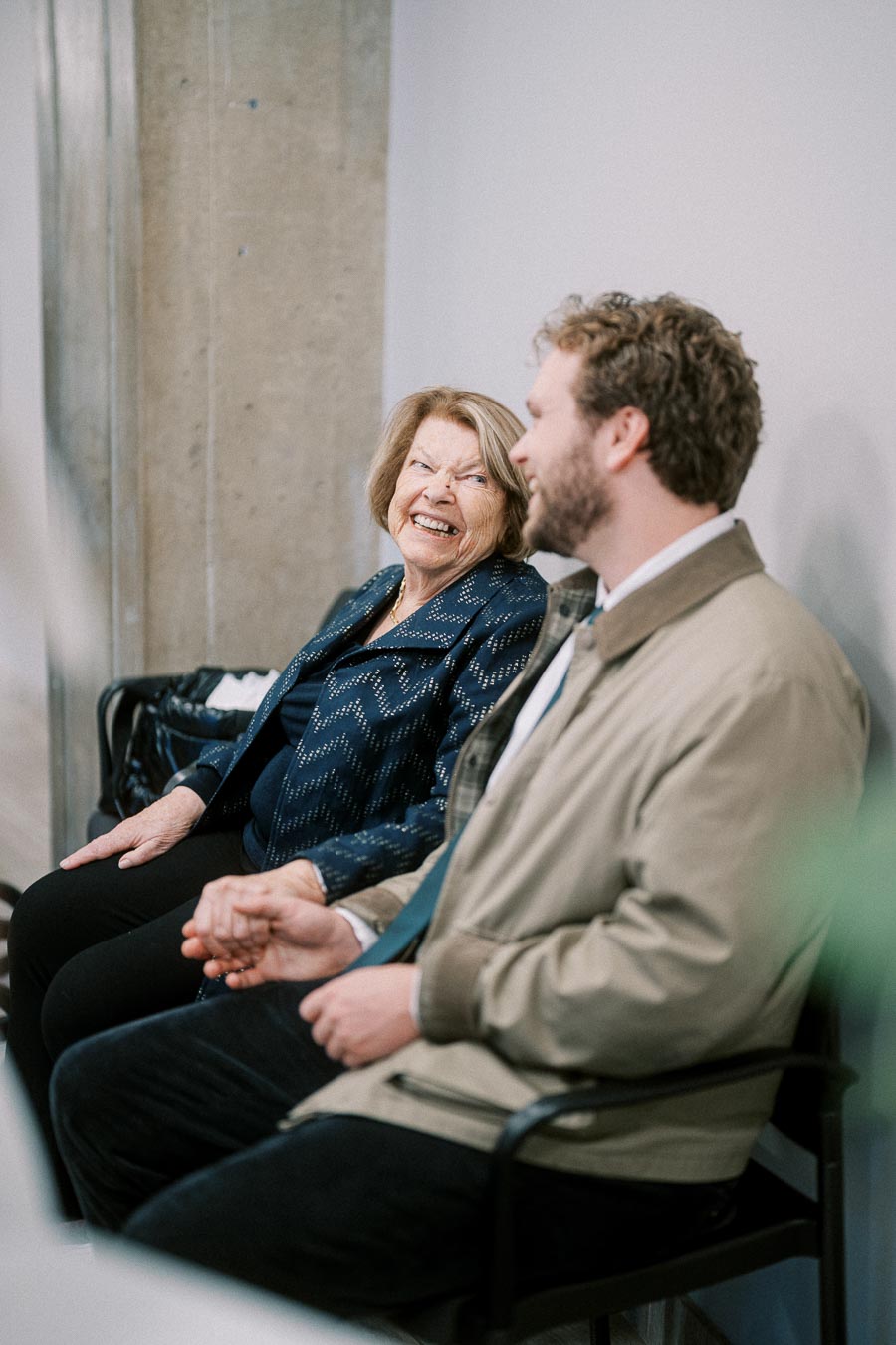 Senior woman and younger man smiling and holding hands while seated indoors, showcasing a warm, joyful moment.