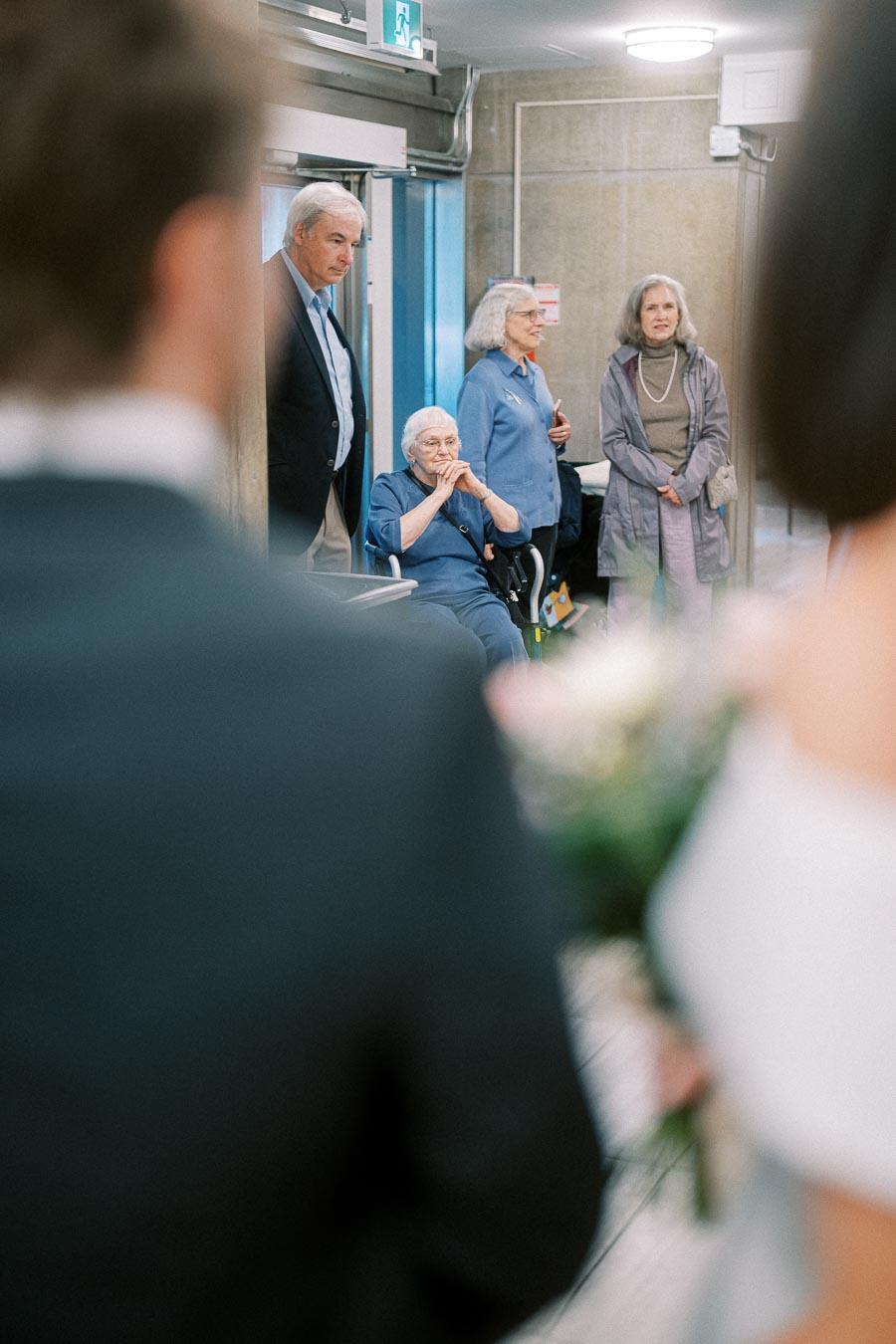 A group of people in a hallway, with one seated in a wheelchair, another in a suit, and two standing women, watching a couple in the foreground during a special event.