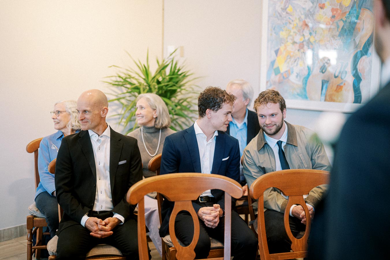 A group of people dressed in formal attire sitting on wooden chairs, engaging in conversation and smiling in a warmly lit room with plants and colorful artwork on the wall.