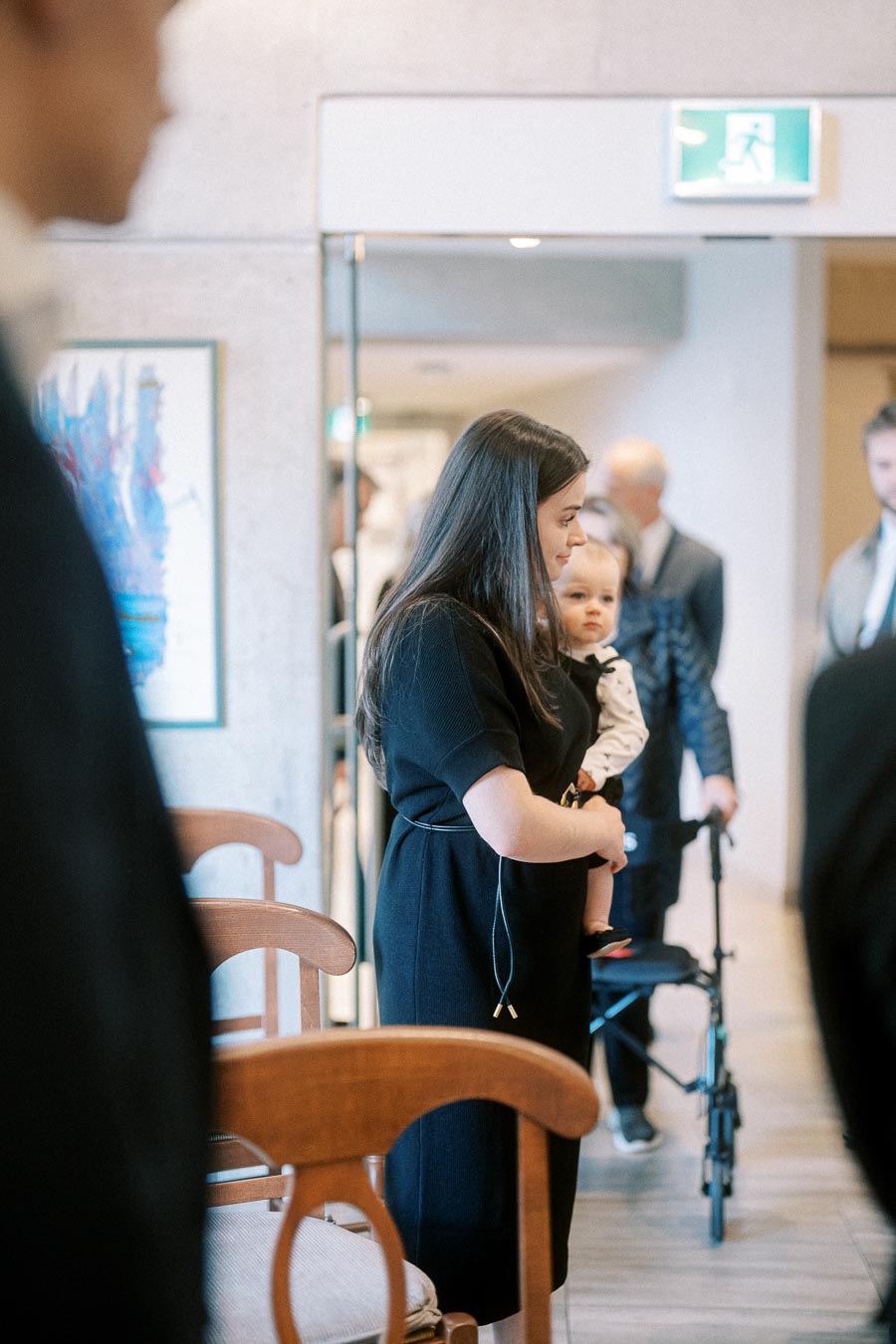 A woman in a black dress holding a baby, standing in an elegantly decorated room with people in the background, conveying a calm and intimate atmosphere.