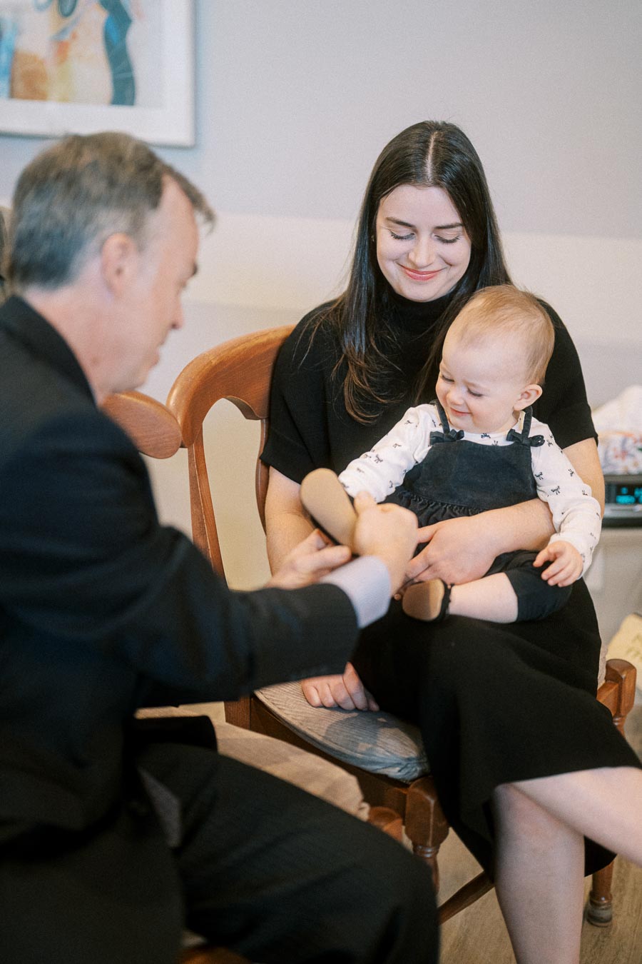 A mother holding her happy baby on her lap while a man in a suit helps put on the baby's shoes, in a cozy home setting.