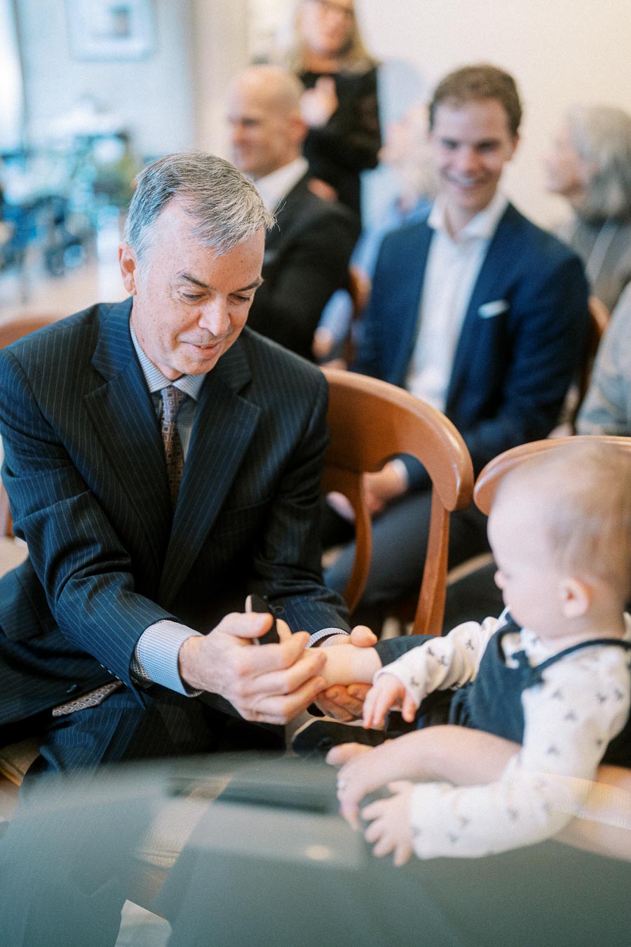 Elderly man in a suit playing with a baby at an indoor gathering, people in the background smiling and engaged.