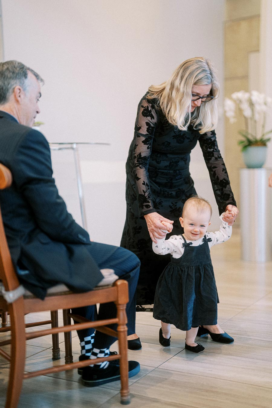 A toddler in a black dress takes assisted steps, guided by an adult woman in a formal outfit, while a seated man watches, creating a joyful family moment indoors.