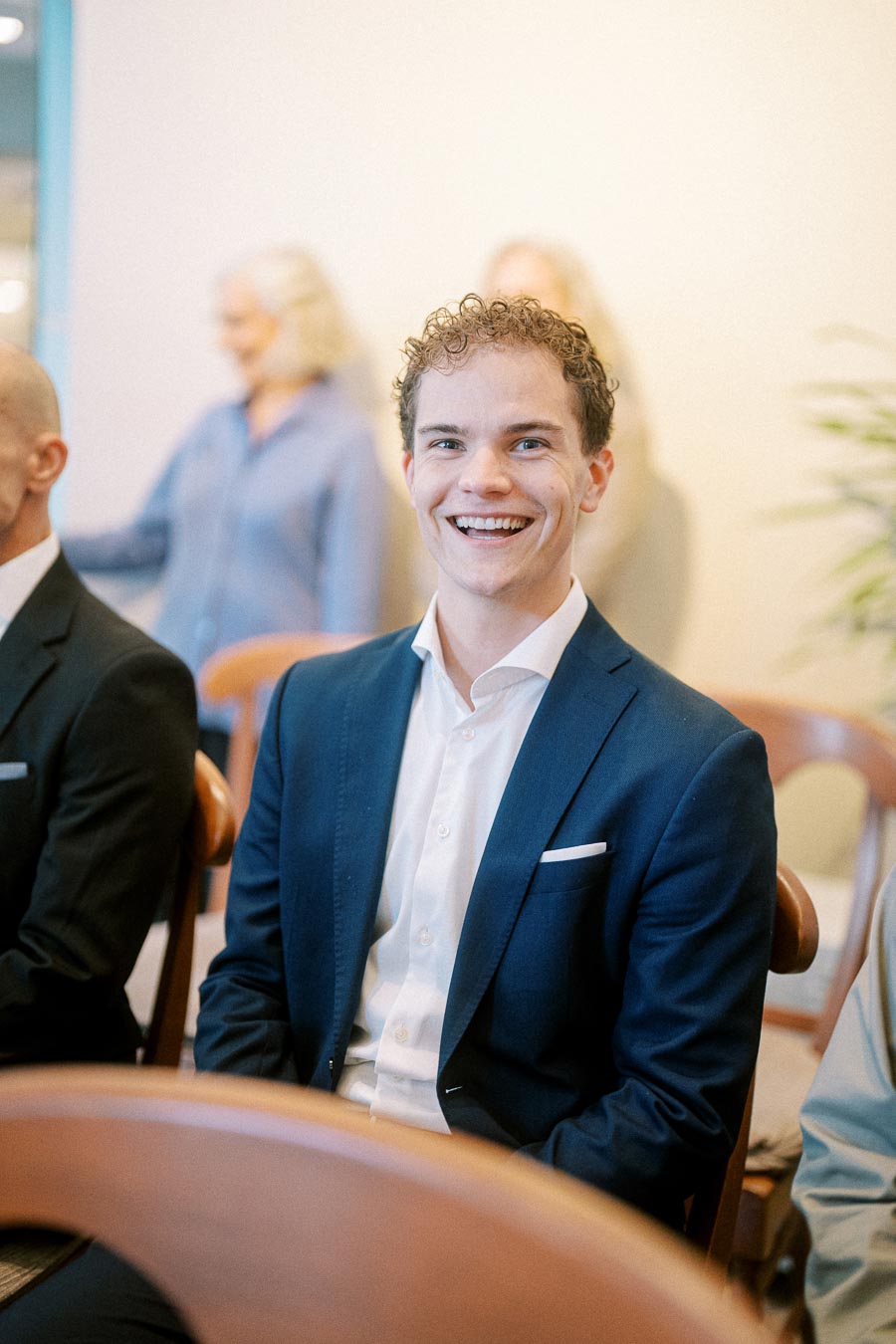 Smiling man in a navy blue suit sitting on a wooden chair in a bright room, with blurred people in the background.