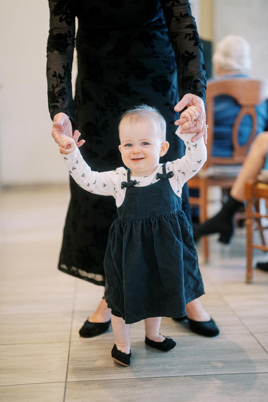A smiling toddler in a white shirt with small patterns and a dark dress being held upright by an adult, learning to walk indoors.