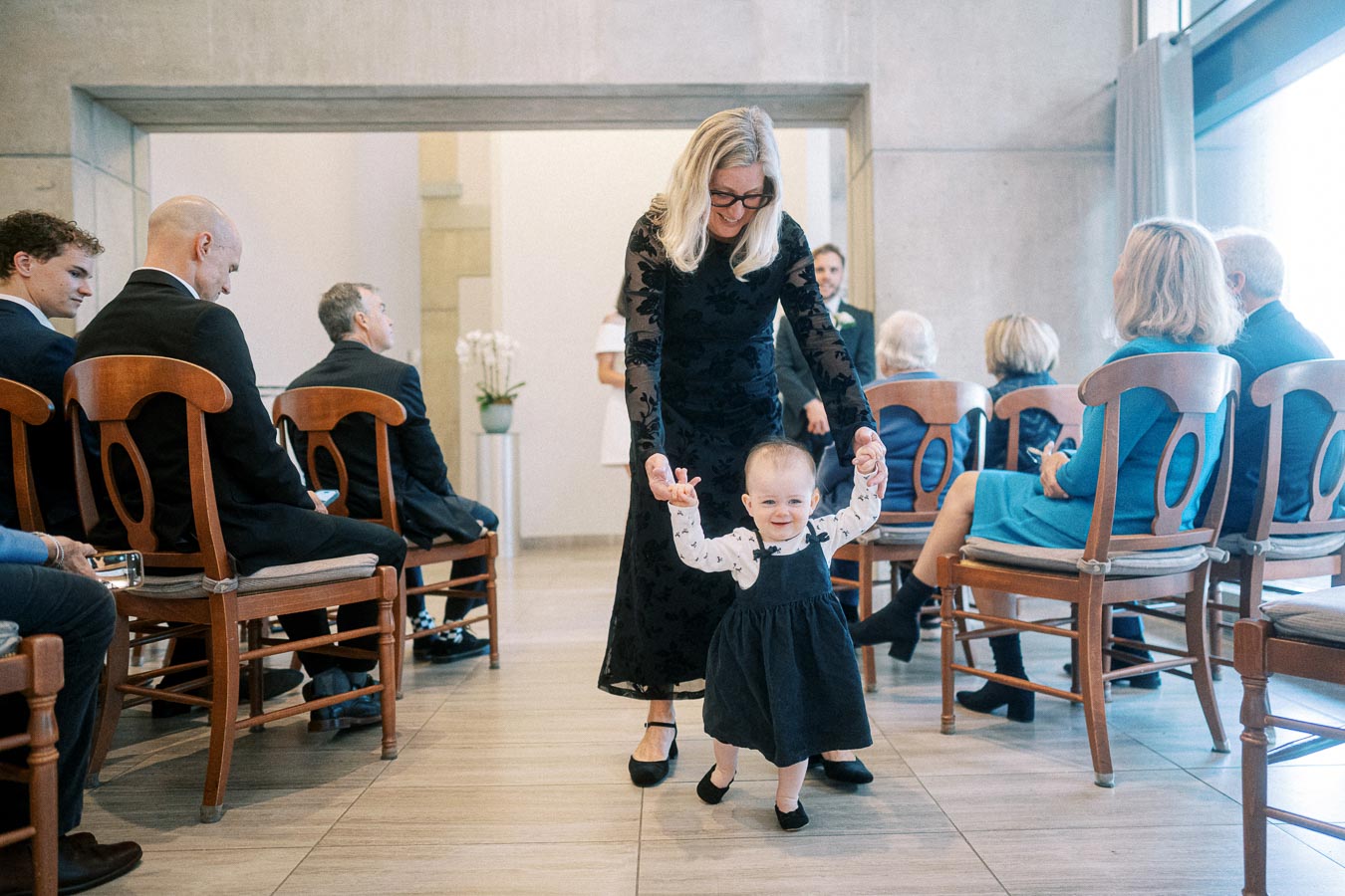 A young child wearing a black dress walks holding hands with a woman in a black dress, surrounded by seated guests at an indoor event.
