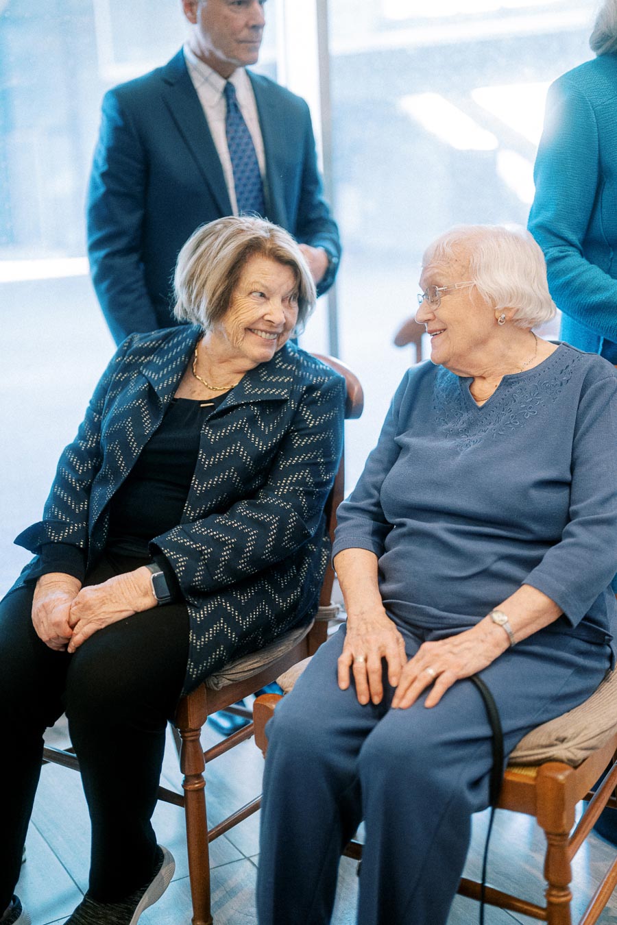 Elderly women sitting and smiling at each other, surrounded by other people, in a social setting.