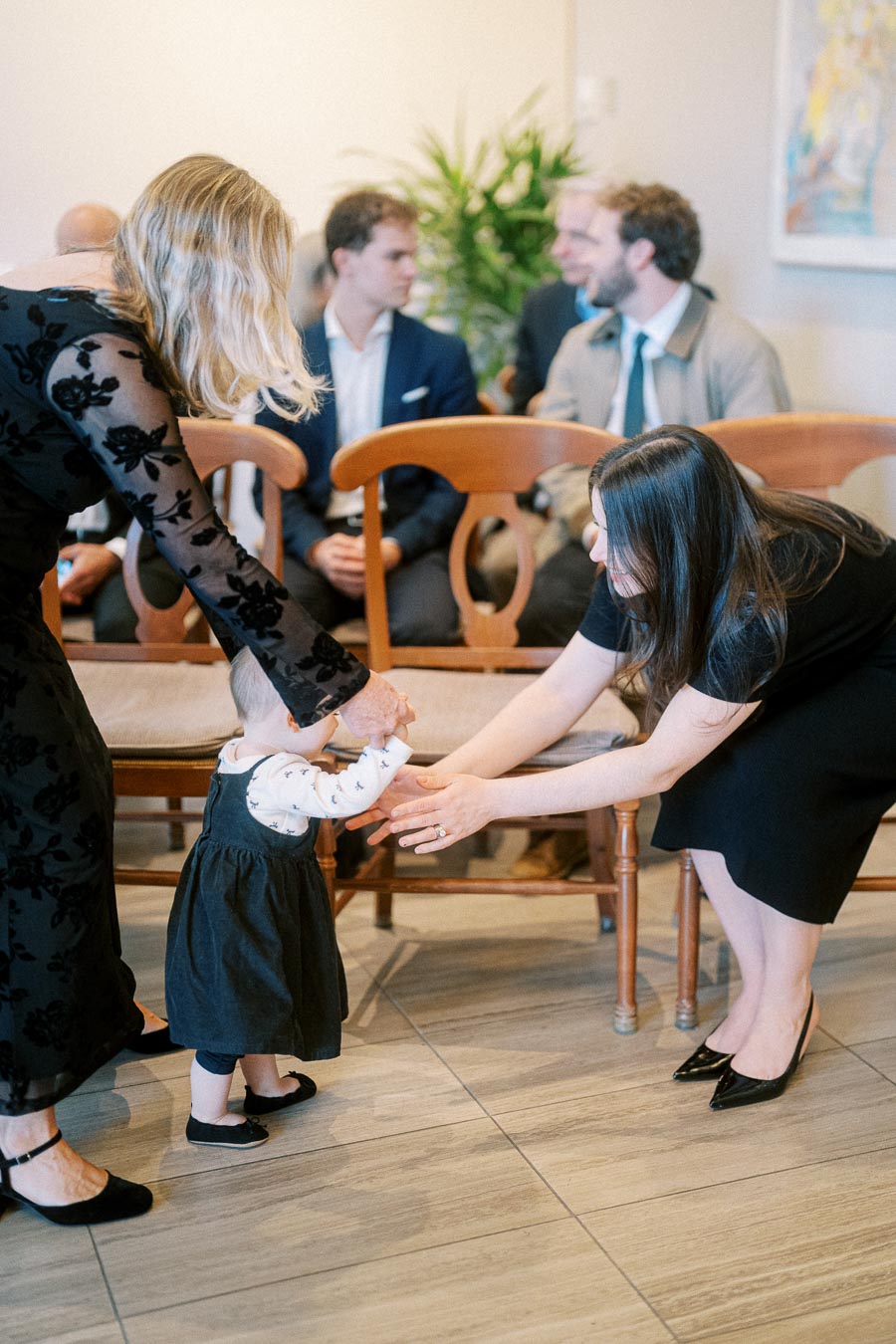 A toddler in a black dress is lovingly supported by two adults, one on each side, in a warmly lit room with chairs and people in the background.