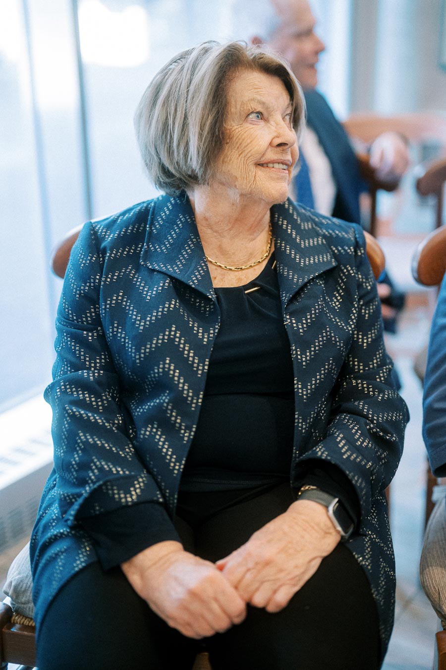 Elderly woman sitting at a gathering, wearing a patterned navy jacket and black top, smiling and looking to the side.
