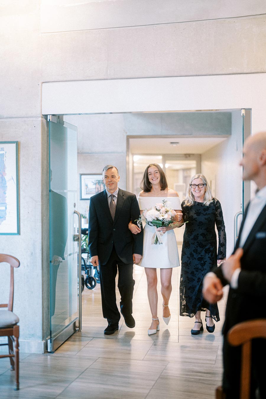 Bride walking with parents down the aisle at a wedding ceremony, wearing a white dress and holding a bouquet, while smiling joyfully.