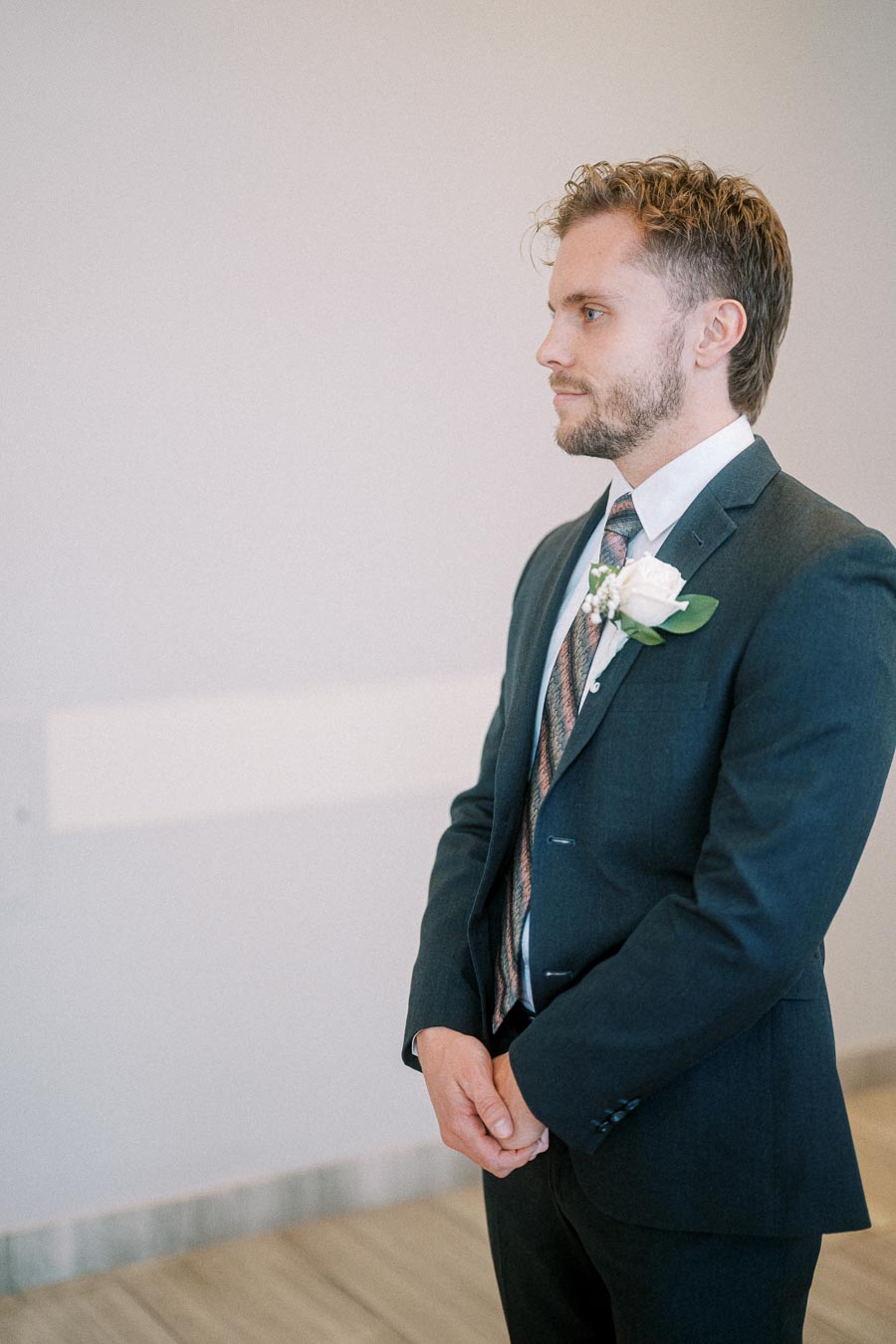A groom in a dark suit adorned with a floral boutonniere, standing in profile, looking composed and ready for his wedding ceremony.