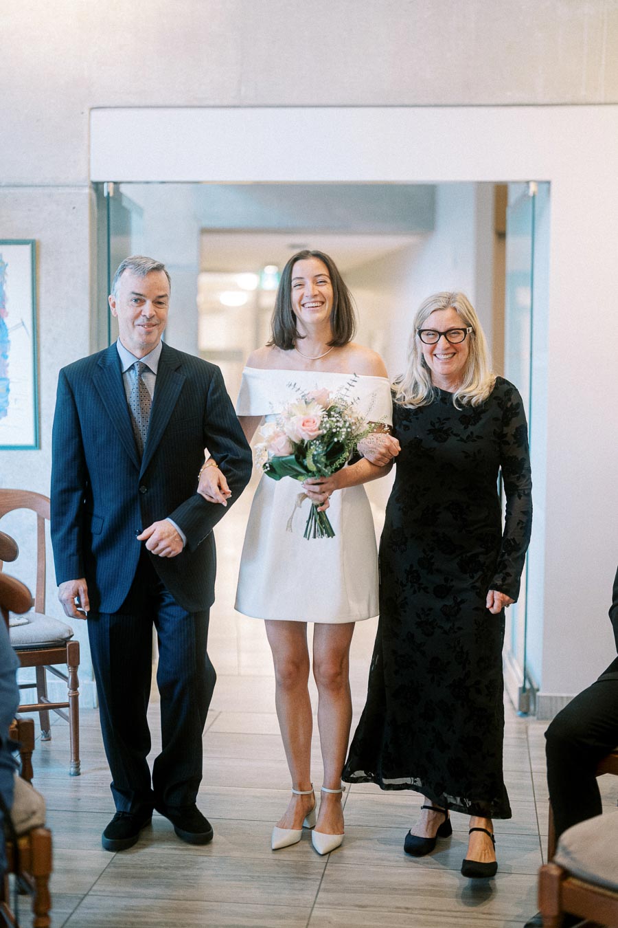 A joyful bride in a white dress holding a bouquet, walking down the aisle with her parents at a wedding ceremony.