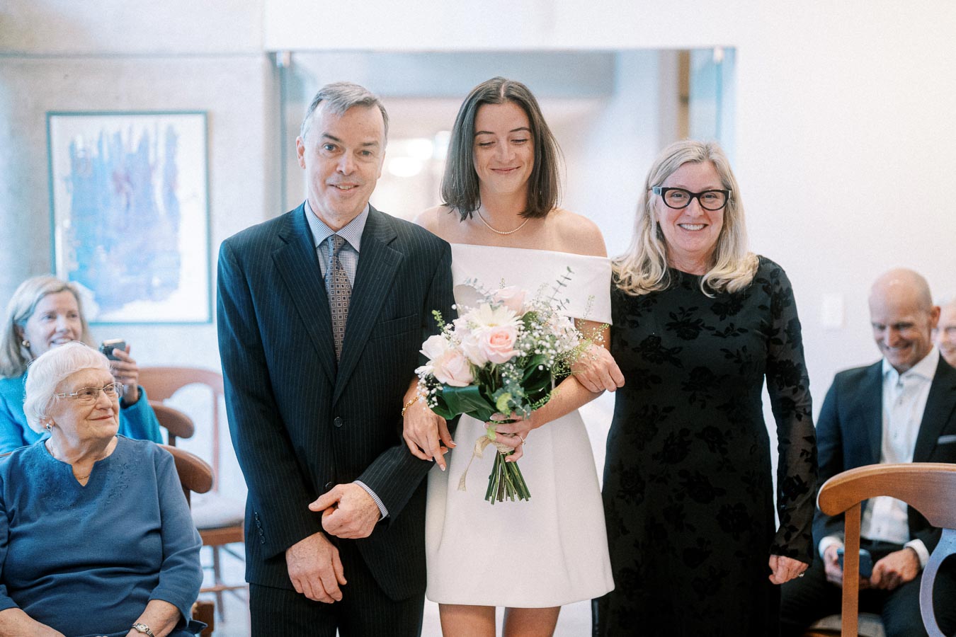 A bride in an elegant white dress holding a bouquet of pink and white roses, walking down the aisle arm-in-arm with two smiling companions in formal attire during a wedding ceremony, with guests seated in the background.