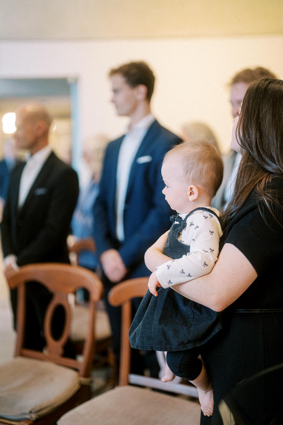 A mother holding her baby at a formal event, surrounded by people in suits, with wooden chairs in the background.