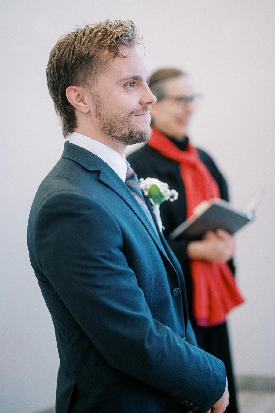 A man in a blue suit stands, smiling slightly, with a boutonniere on his lapel, in what appears to be a formal setting. Another person in the background is holding a book, wearing a red scarf.
