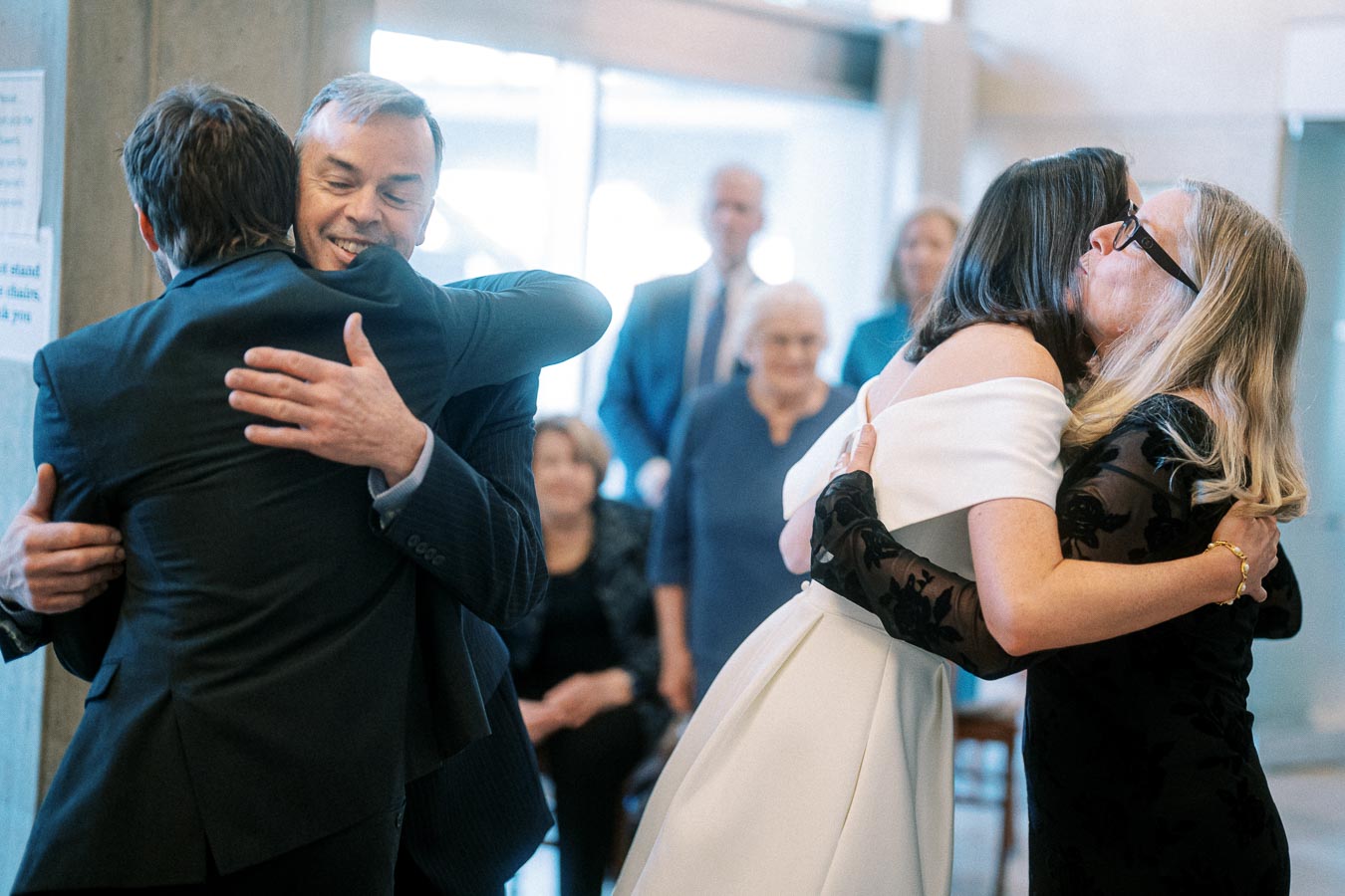 A joyful wedding scene where two couples warmly embrace each other in celebration, surrounded by elegantly dressed guests in a bright and cheerful atmosphere.