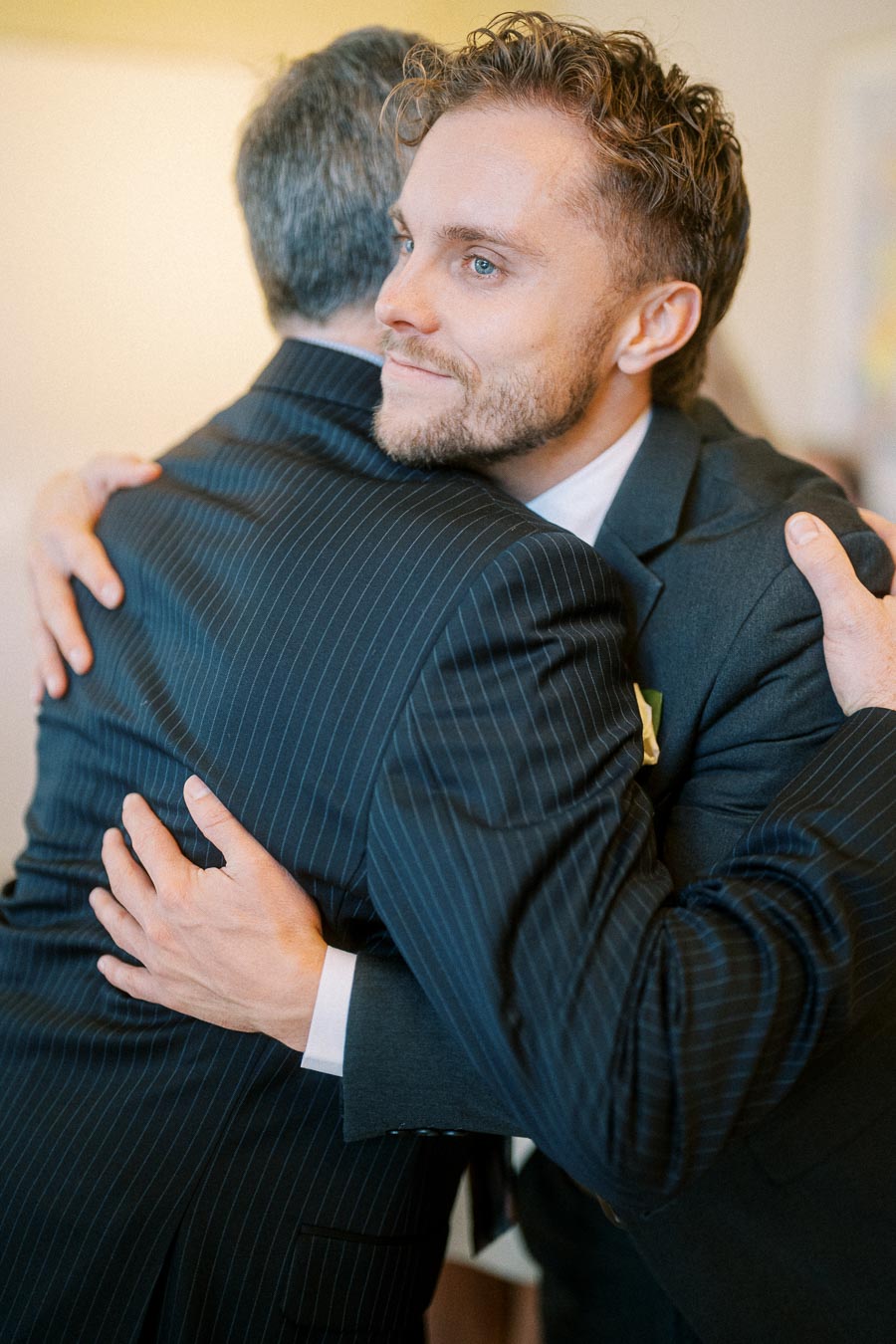 A man with curly hair and a beard hugs another man wearing a pinstripe suit.