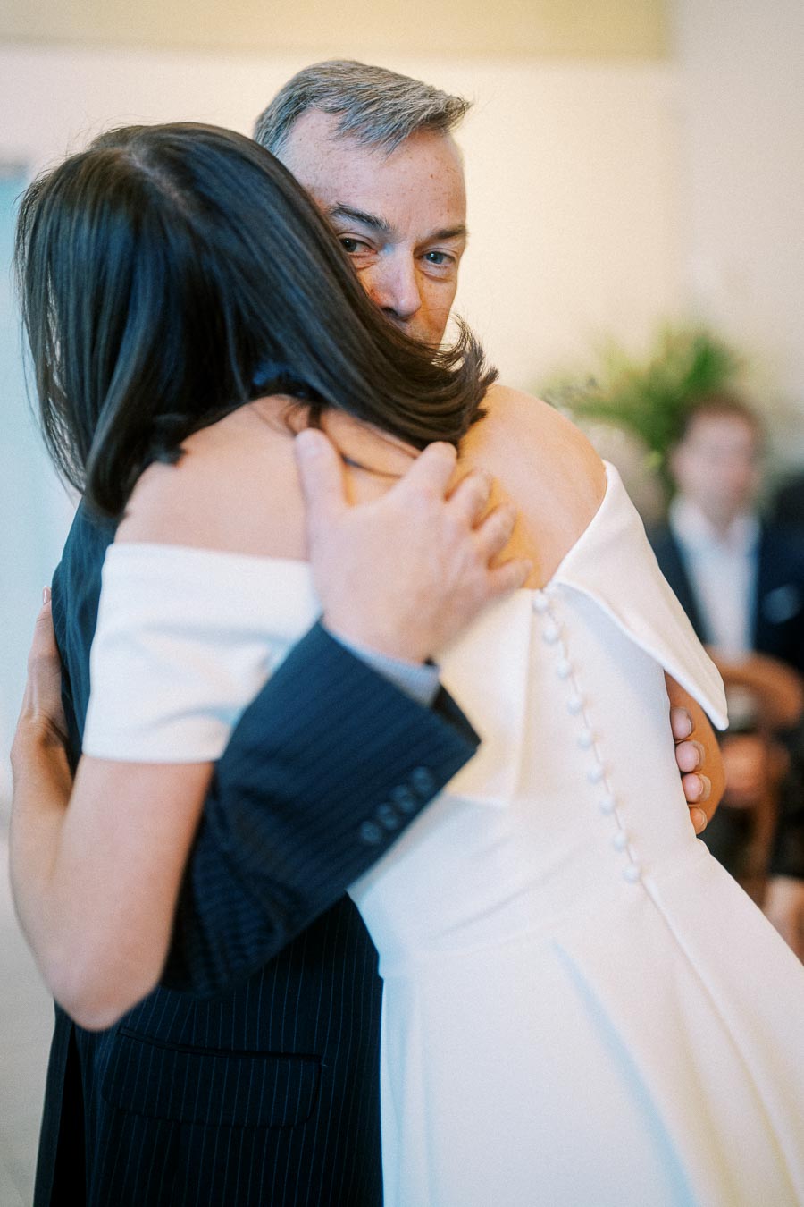 Two people embracing at a wedding, with the bride wearing an elegant white dress featuring button details, and the groom in a dark suit. The background has a soft, blurred ambience with greenery and guests, highlighting an intimate celebratory moment.