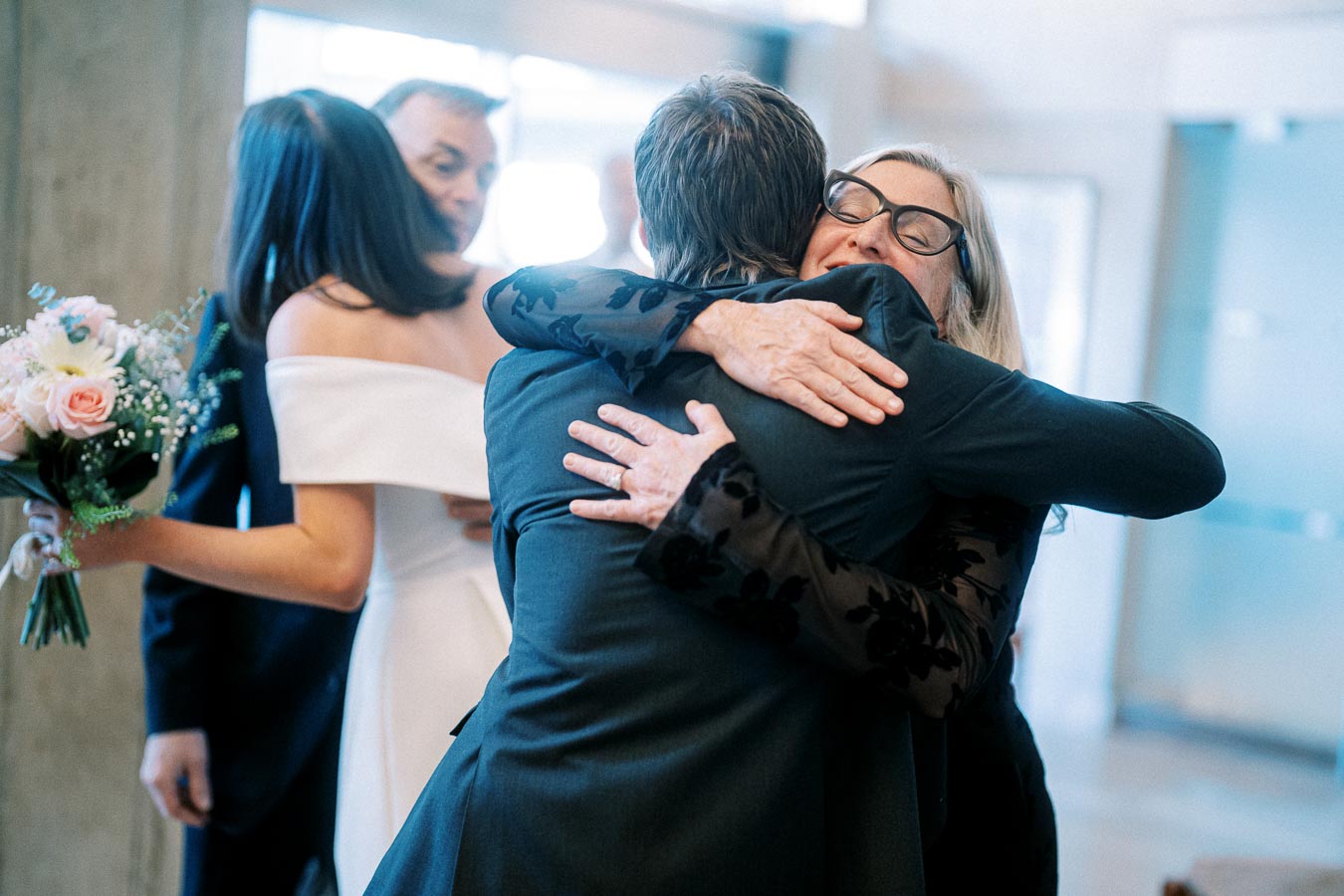 A joyful wedding scene showing a bride in an off-the-shoulder dress holding a bouquet while guests warmly embrace, celebrating love and unity.