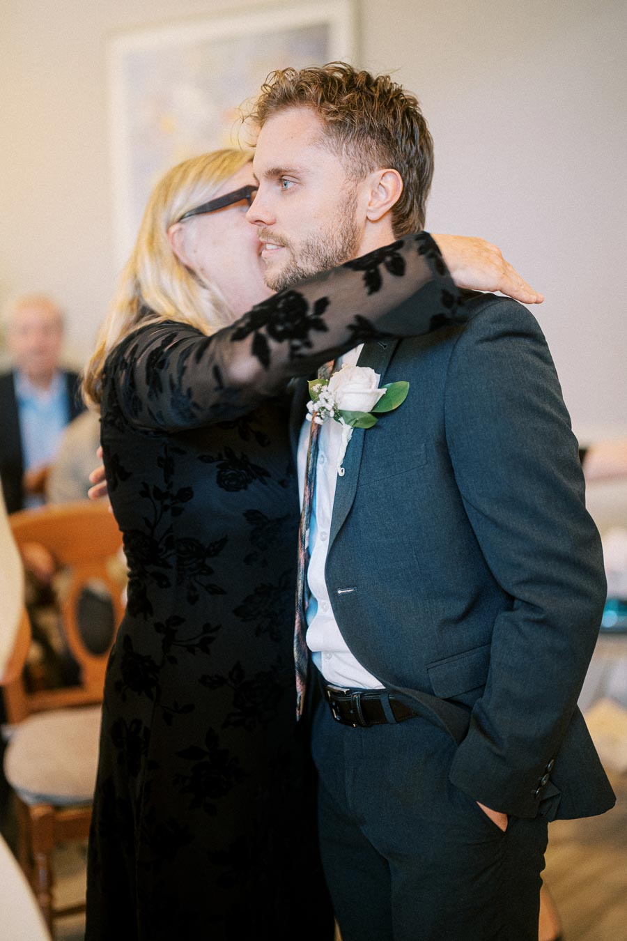 A man in a suit receives an affectionate hug from a woman in a black floral dress, during a special gathering or event indoors.