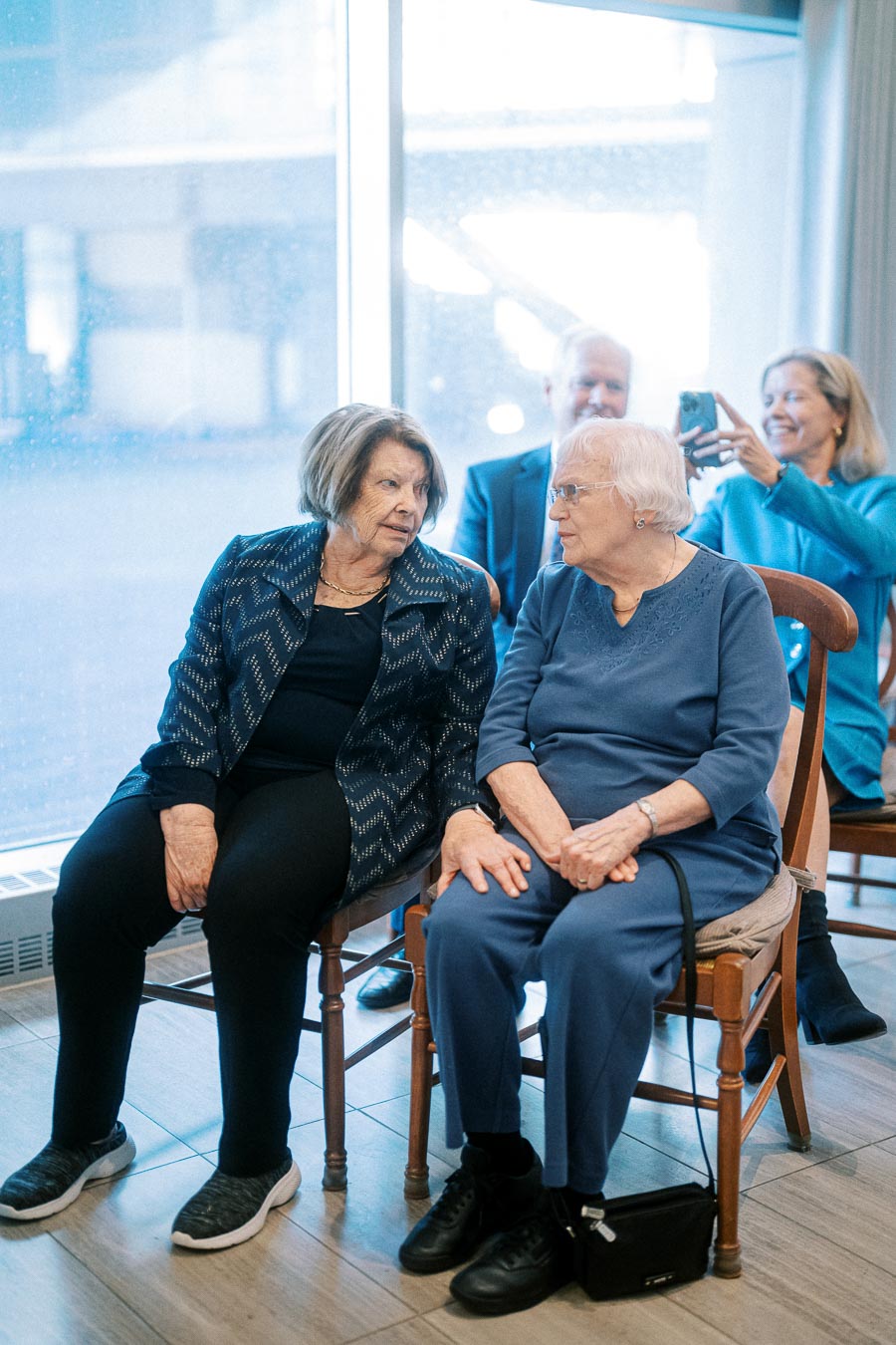 Two elderly women sitting and conversing at a gathering, with a man taking a photo in the background.