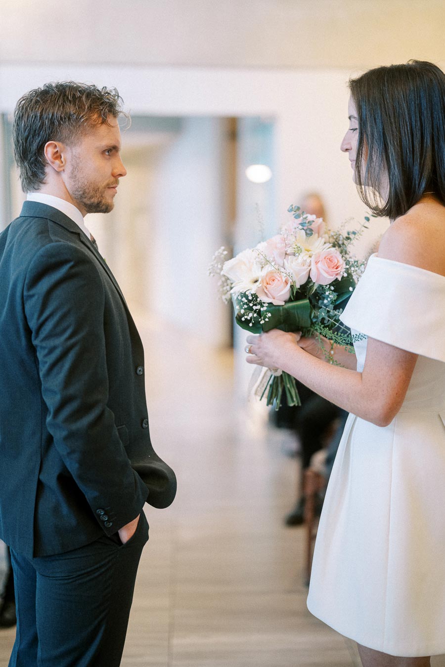 A bride holding a pink and white bouquet faces a groom in a suit during an intimate indoor wedding ceremony.