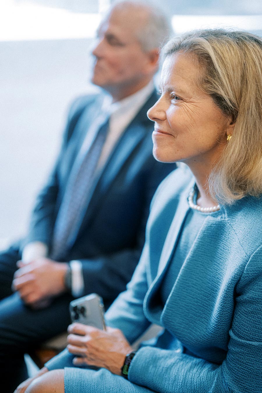 Two professionals attentively listening during a business meeting, wearing formal attire and sitting side by side, with focus on the woman holding a smartphone.