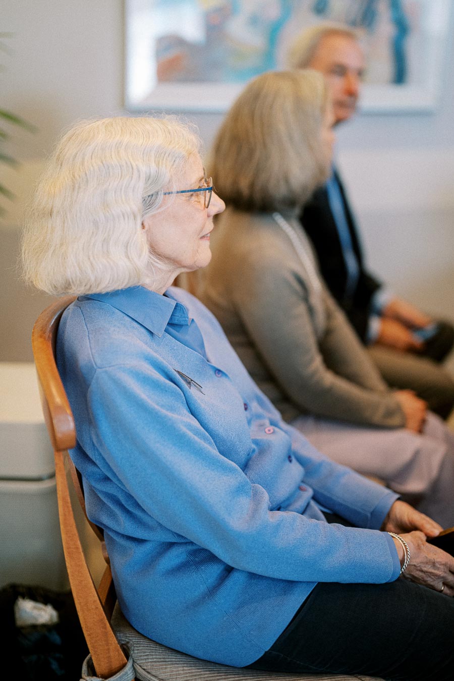 Elderly woman in a blue sweater attending a meeting, seated in a row with others in a calm, professional setting.