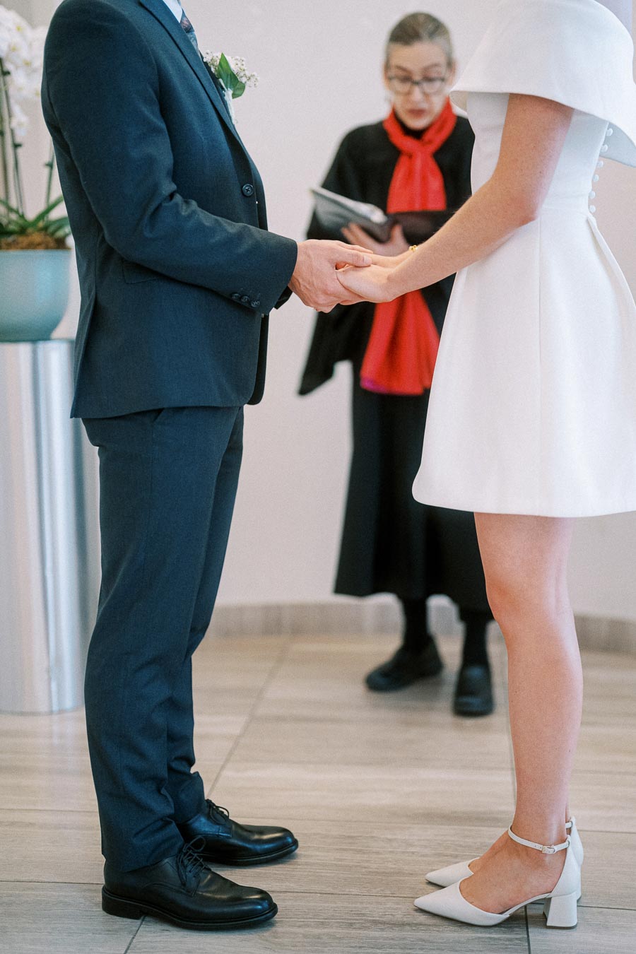 A couple holding hands during their wedding ceremony, with a celebrant officiating in the background. The groom wears a dark suit, and the bride is in a white dress and heels, standing in an elegantly decorated venue.