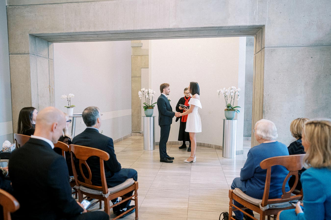 A couple exchanges vows during an intimate indoor wedding ceremony, with guests seated nearby. The bride wears a white dress, and the groom is in a suit. The minimalist setting is decorated with orchids, enhancing the elegant atmosphere.