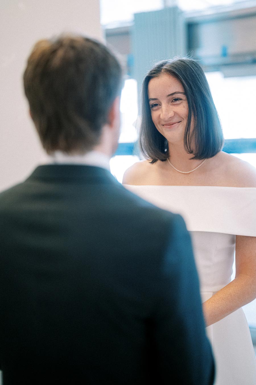 A bride in a white wedding dress smiling at her groom during their wedding ceremony inside a bright, modern venue.