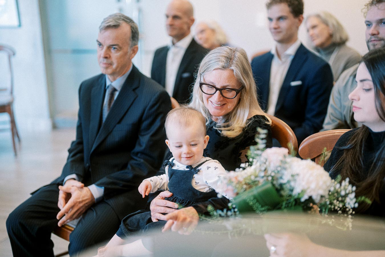 Grandmother holding baby with family sitting in formal attire at a ceremony, focus on baby and floral arrangement.