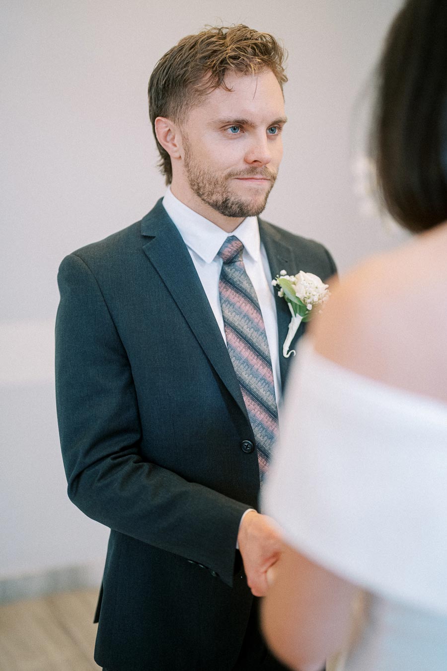 A groom in a blue suit and patterned tie stands facing his bride during an intimate wedding ceremony, with a white floral boutonniere on his lapel.