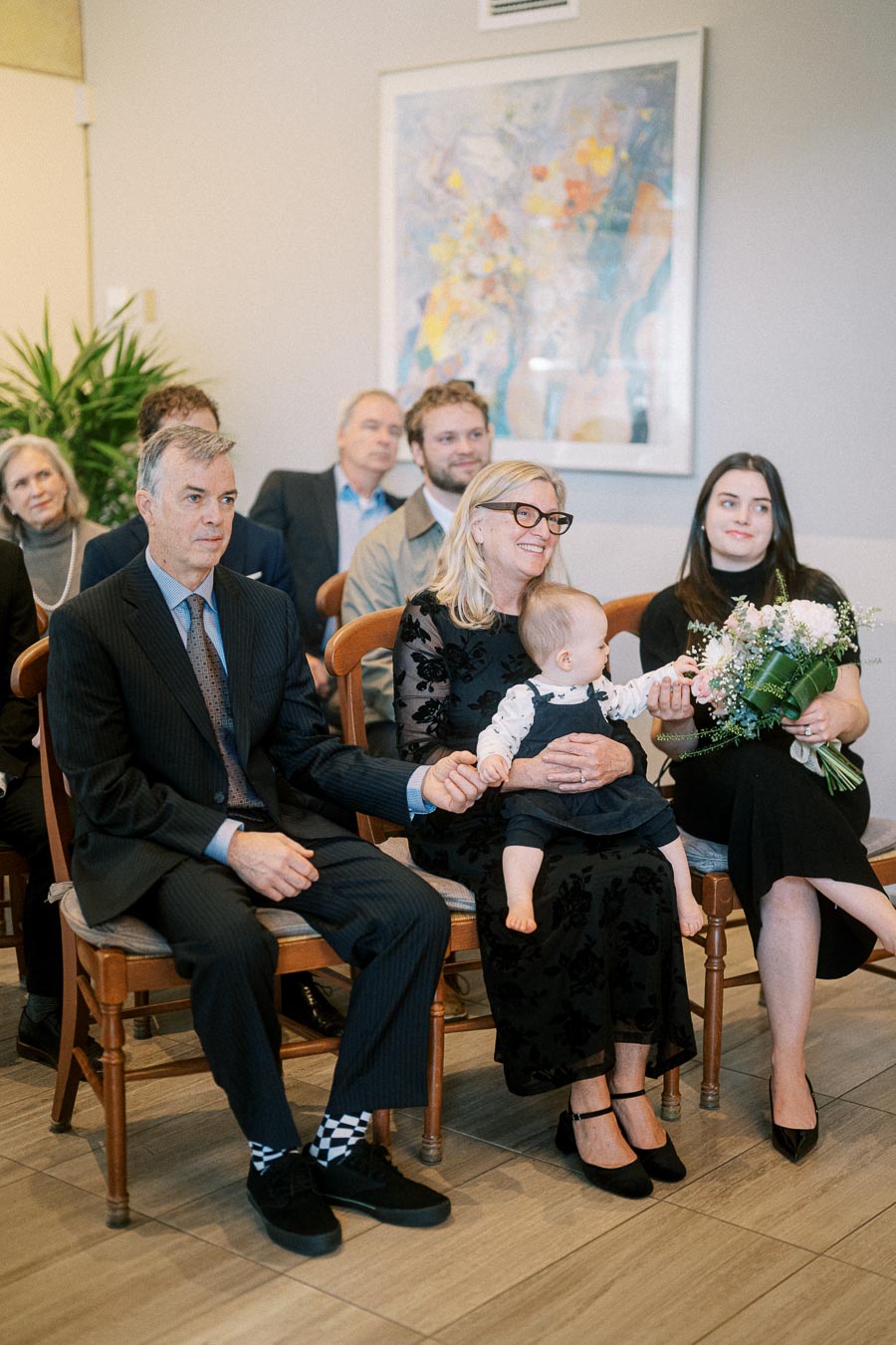 Family attending a formal gathering, featuring a man in a suit, a woman holding a baby, and another woman with a bouquet, all seated in a warmly lit room with framed artwork in the background.