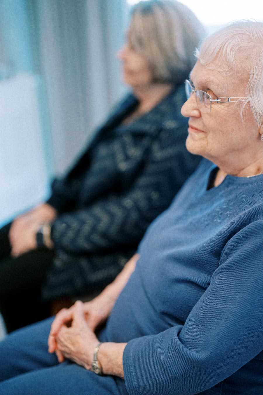 Elderly woman with glasses sitting and listening attentively in a room, wearing a blue outfit and a watch on her wrist.