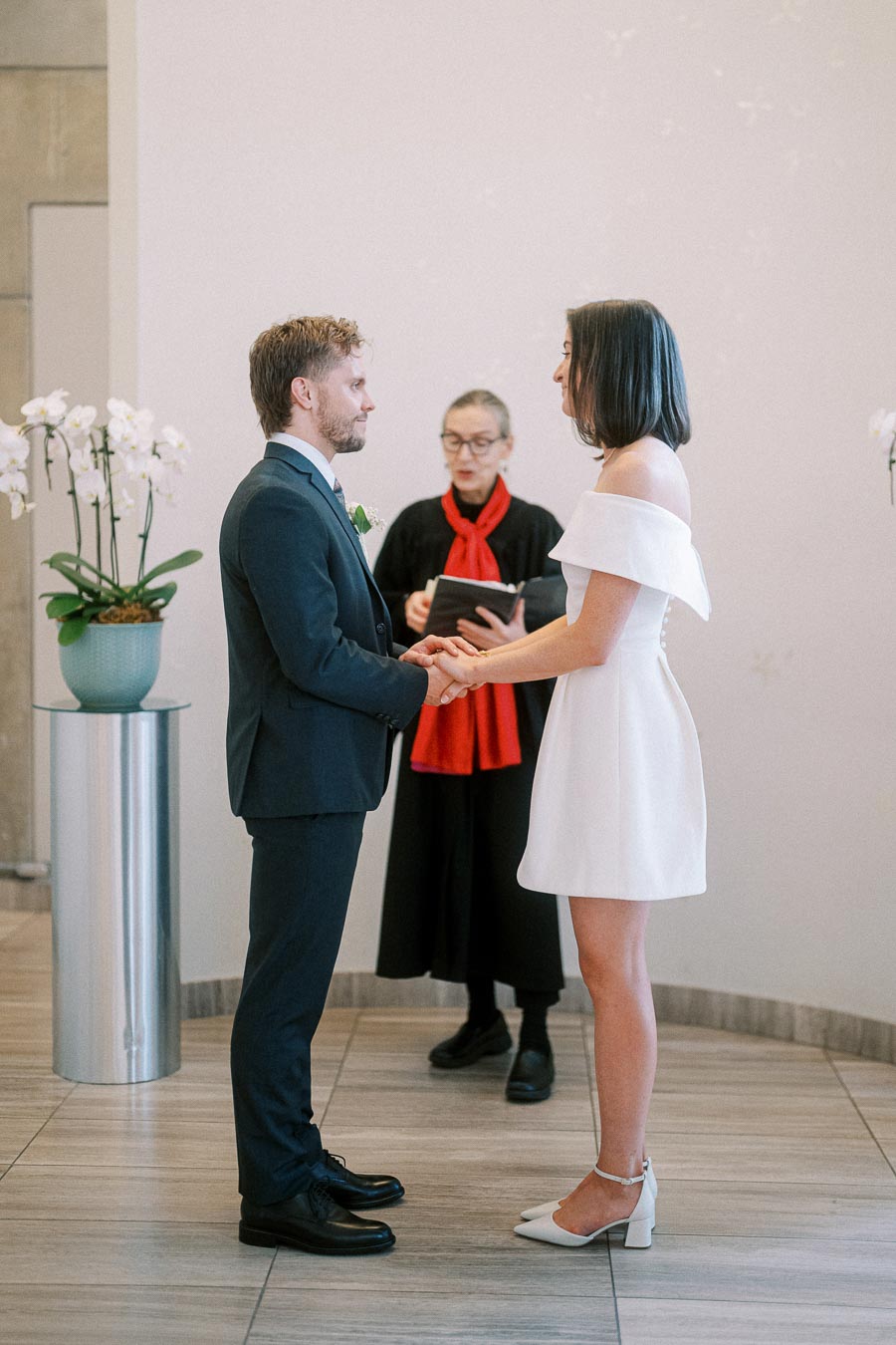 A couple exchanging vows during a modern indoor wedding ceremony; the bride is in a white dress and the groom in a navy suit, with an officiant in the background and orchid arrangements nearby.