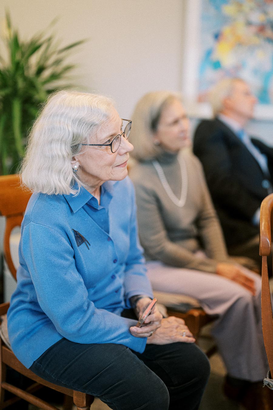 Elderly woman with gray hair and glasses attentively listening during a community meeting, while seated beside other participants in a warmly lit room.