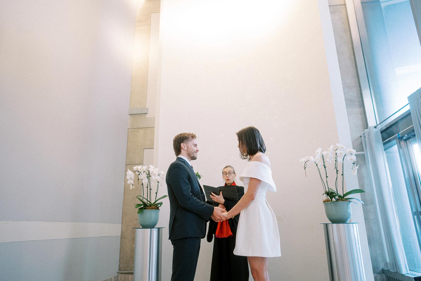 A couple exchanges vows during a modern indoor wedding ceremony, with a minimalist decor featuring tall potted white orchids on either side, and a dressed officiant holding a black folder in the background.