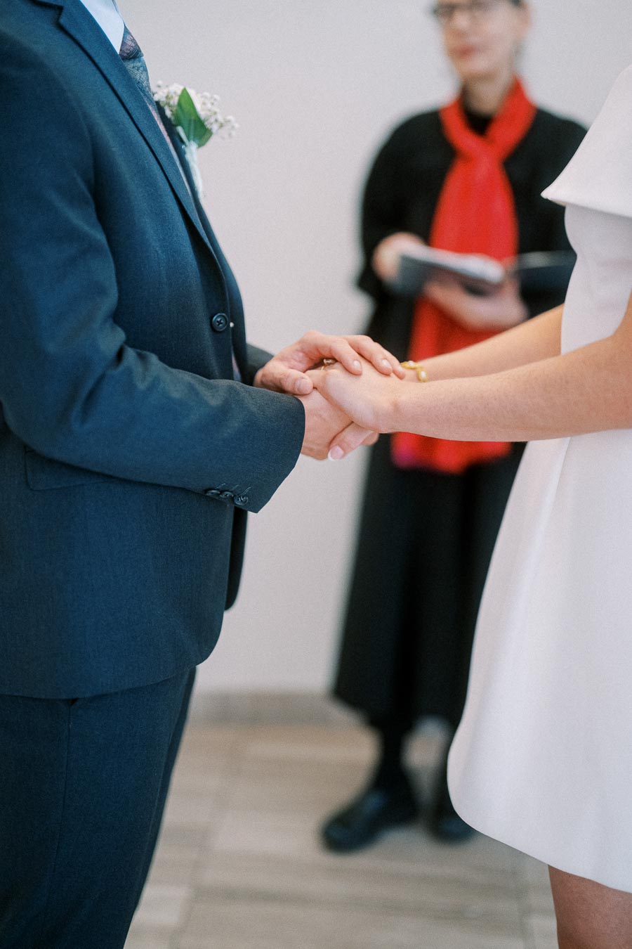 A couple holding hands during a wedding ceremony, with an officiant in the background.