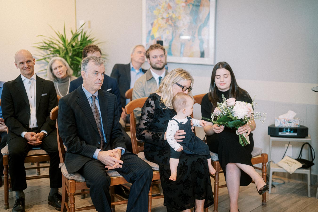 A group of people seated at an indoor event, featuring a woman holding a baby and another woman holding a bouquet of flowers. The attendees appear to be focused and attentive, with a large potted plant and abstract artwork visible in the background.