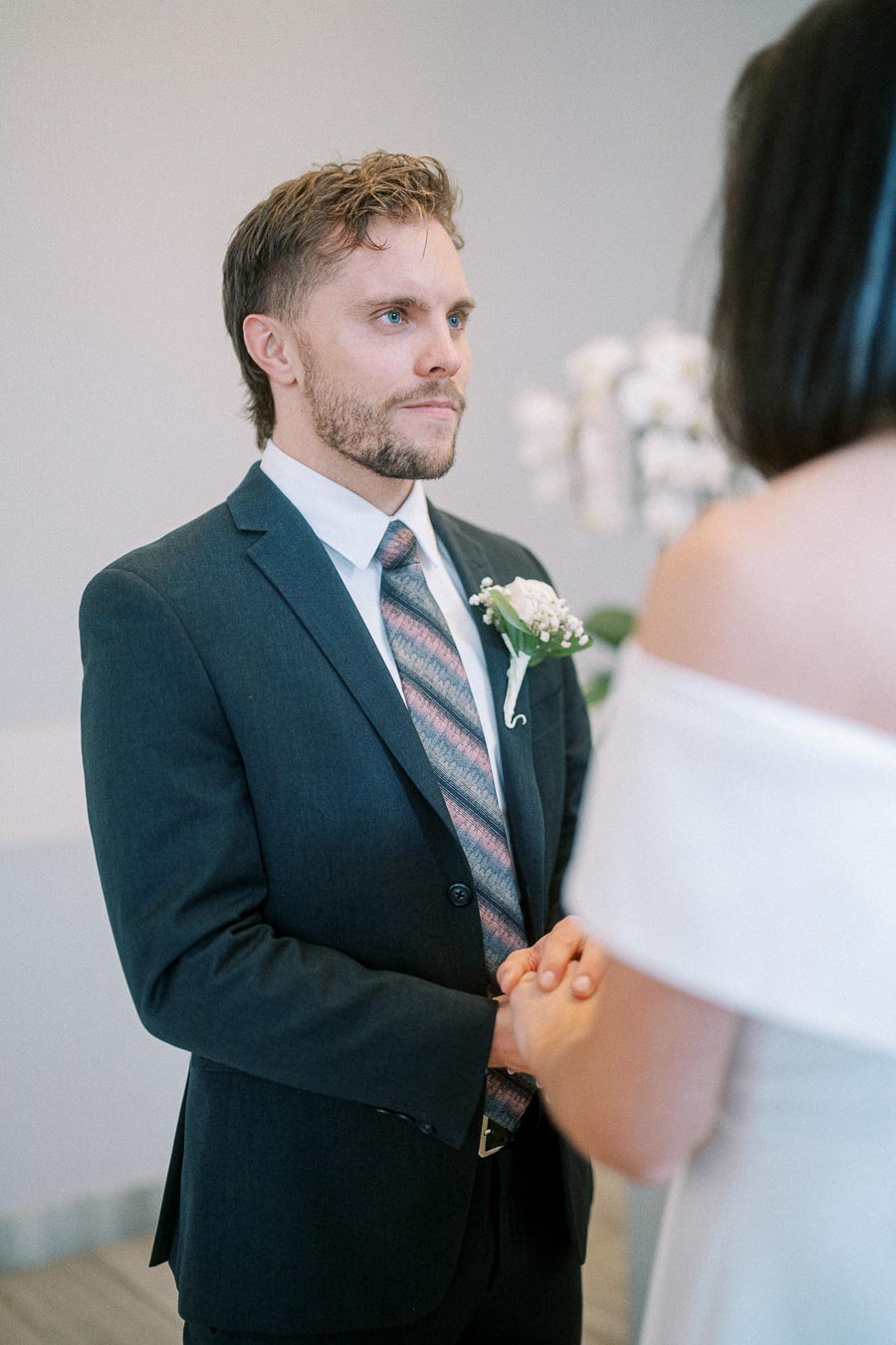 A groom in a dark suit and tie holds hands with a bride during an intimate wedding ceremony.