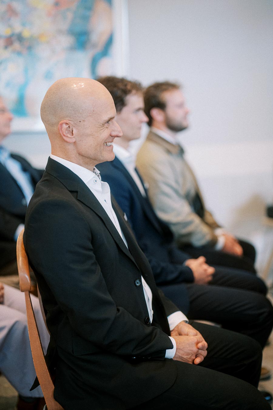 A group of professionals in suits attentively seated in a seminar room, engaged in a presentation or meeting, with a colorful abstract painting in the background.