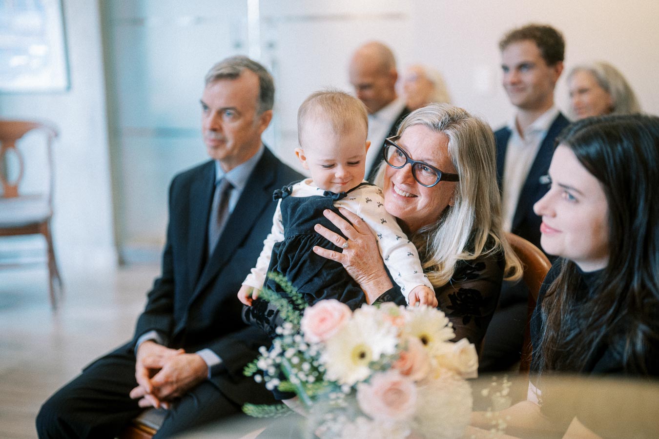 A joyful family gathering with a smiling elderly woman holding a baby, surrounded by attentive adults, and a bouquet of flowers in the foreground.