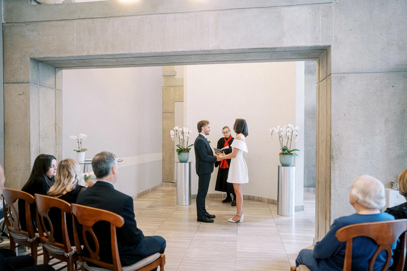 A couple exchanging vows in a modern indoor wedding ceremony, with guests seated and an officiant in the background.