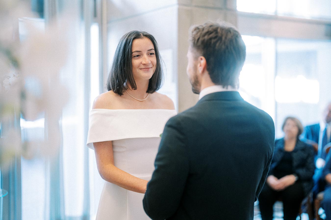 A bride and groom exchanging vows during a wedding ceremony, with the bride wearing an off-shoulder white dress and the groom in a suit, indoors with guests seated in the background.